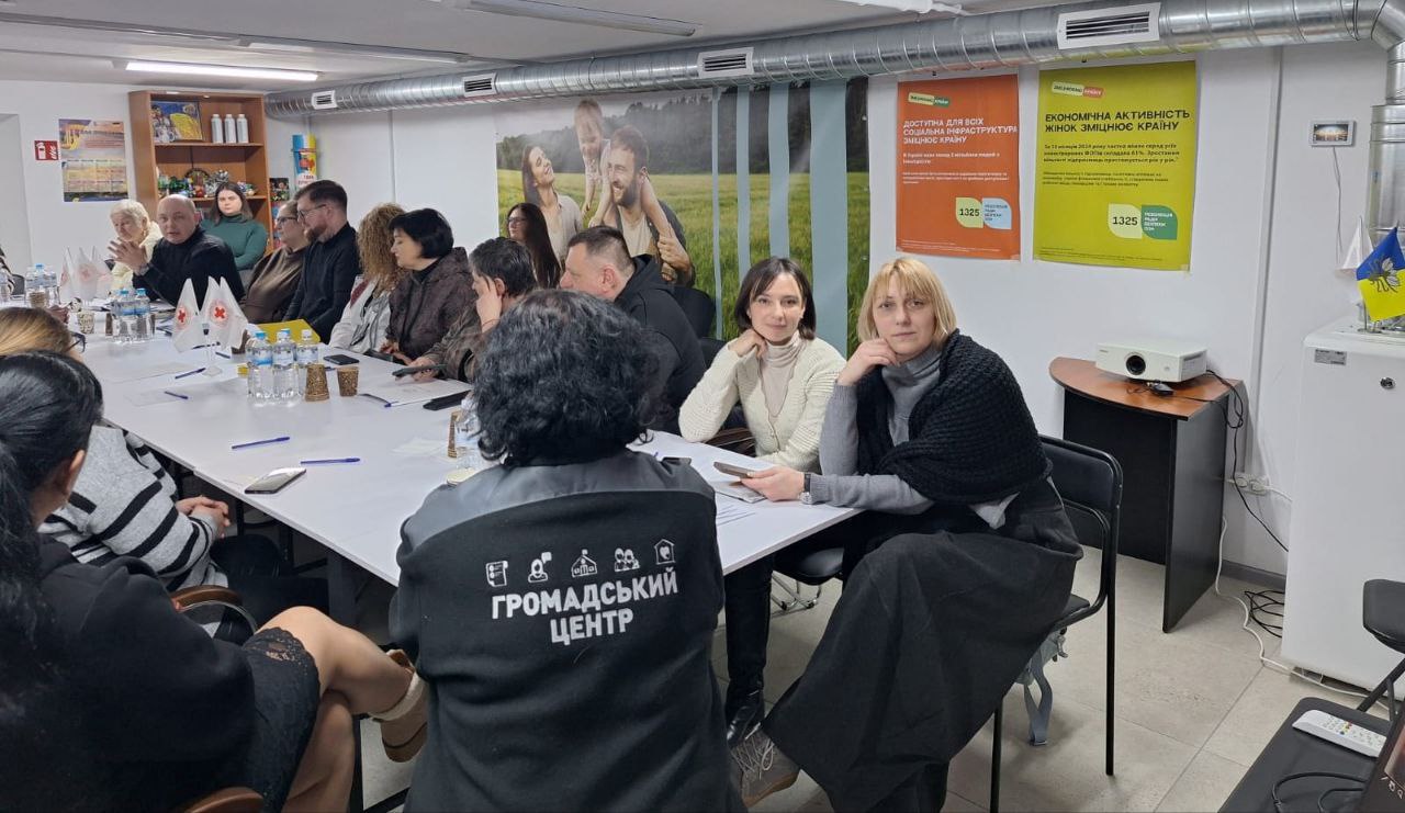 Учасники наради сидять за довгим столом у приміщенні громадського центру. На стінах розміщені інформаційні плакати про соціальну інфраструктуру та економічну активність жінок, а на столі видно маленькі прапорці з логотипом Червоного Хреста.