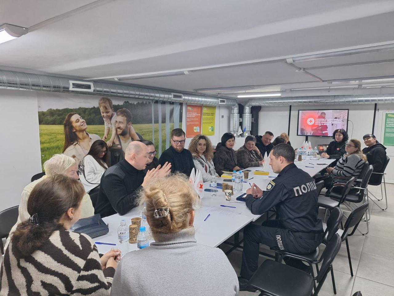 Група людей сидить за великим столом під час робочої зустрічі або наради в офісному приміщенні. Серед присутніх — представник поліції у формі. На столі видно папери, пляшки з водою та символіку Червоного Хреста. На стіні розміщено великий плакат із зображенням усміхненої сім'ї на фоні природи.