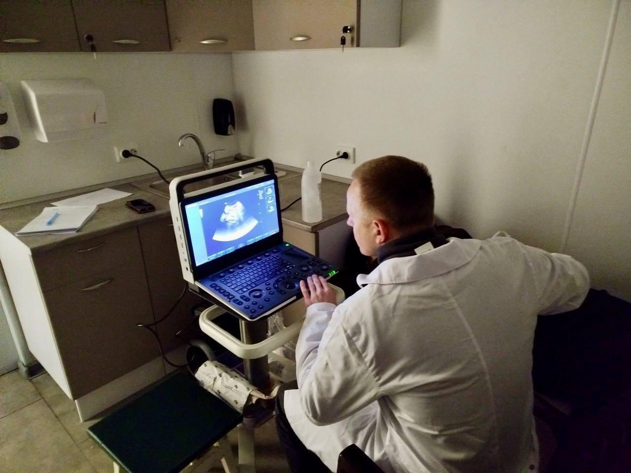 Медичний працівник у білому халаті та захисній масці проводить ультразвукове дослідження пацієнту за допомогою портативного апарату УЗД у кабінеті.