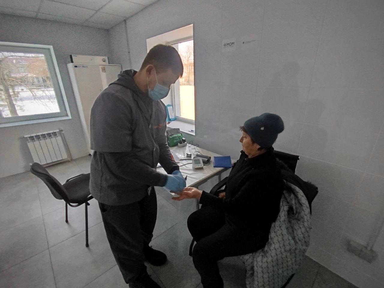 Медичний працівник у захисній масці та рукавичках проводить огляд пацієнтки в облаштованому кабінеті. Спеціаліст тримає руку жінки, ймовірно, для забору крові або вимірювання рівня цукру. На столі поруч видно тонометр та інше медичне приладдя.