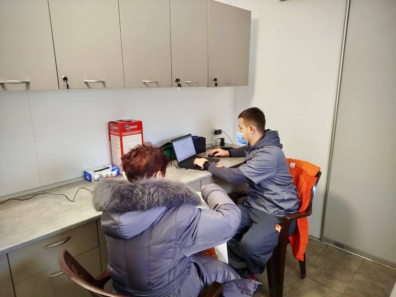 Медичний працівник у захисній масці працює за ноутбуком, вносячи дані під час прийому пацієнтки. Жінка сидить поруч за робочим столом у кабінеті. На фоні видно медичні меблі, контейнер для медичних відходів та витратні матеріали.