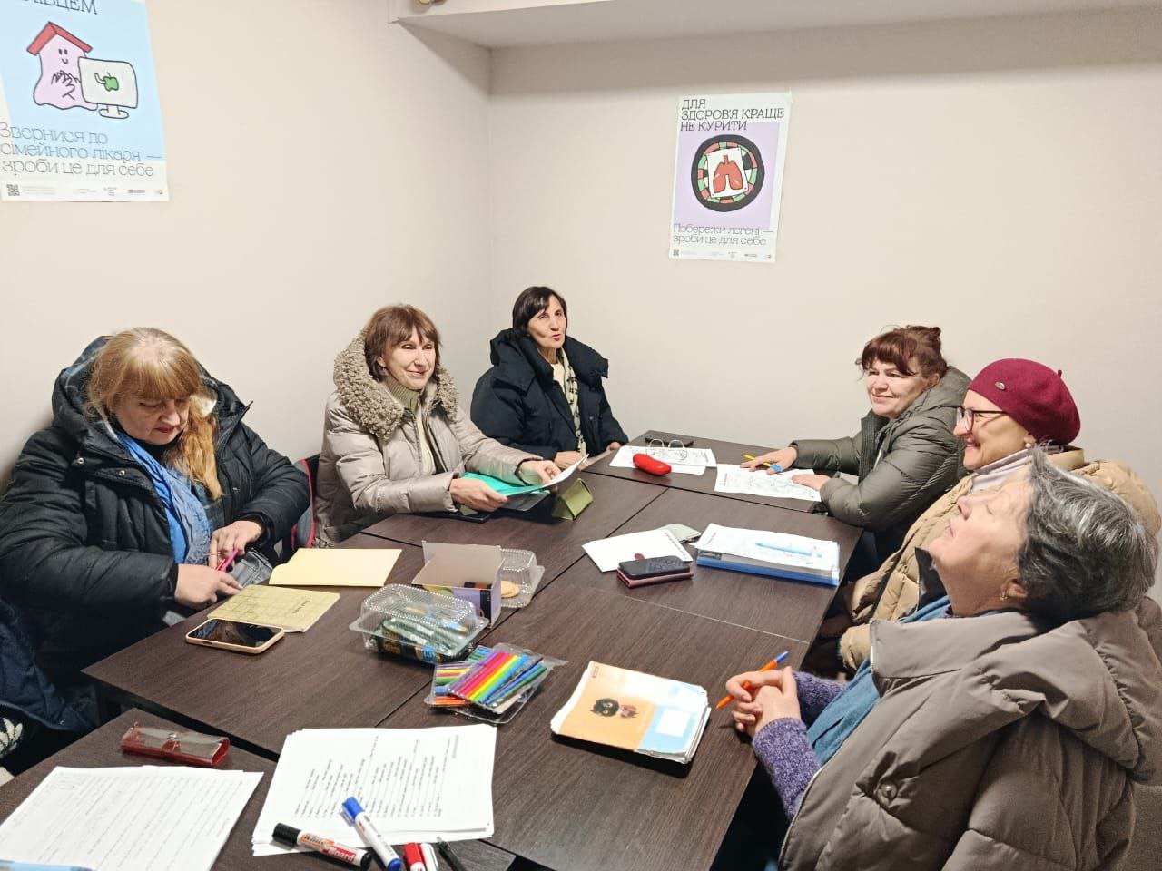 Група жінок, одягнених у верхній одяг сидить за великим столом у навчальному класі. Вони розглядають документи, блокноти та канцелярське приладдя, що лежать перед ними. На стінах кабінету розміщені плакати на медичну тематику українською мовою.