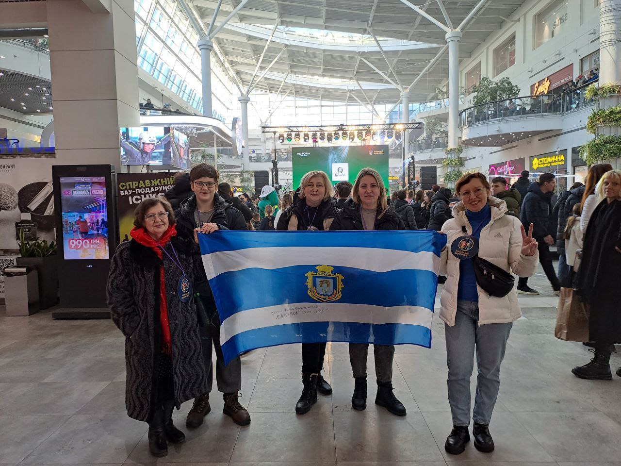 П'ятеро людей стоять у просторому холі торгового центру та тримають розгорнутий синьо-білий прапор із гербом м.Херсона посередині. На задньому плані видно сцену та інтер'єр ТРЦ.