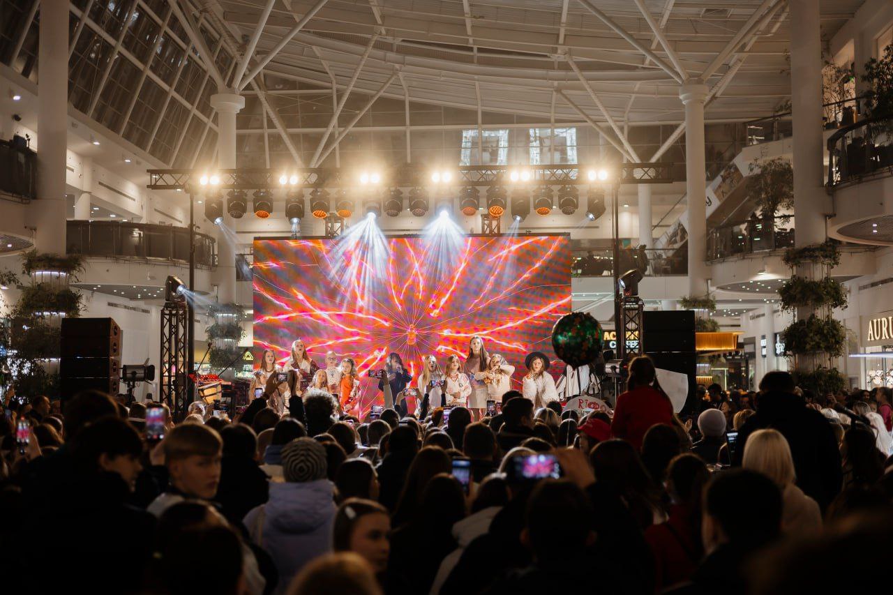 Група молодих виконавців виступає на сцені в центрі великого залу торгового центру. Позаду них працює великий екран із яскравою графікою та професійне освітлення, а навколо зібралося багато глядачів.