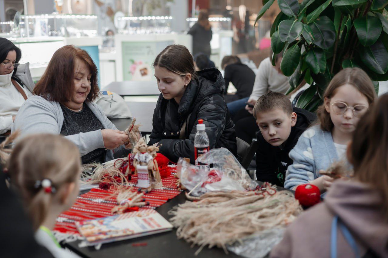 Доросла жінка та кілька дітей сидять за столом, займаючись виготовленням традиційних ляльок-мотанок. На столі лежать нитки, солому та інші матеріали для рукоділля; подія відбувається в публічному місці.