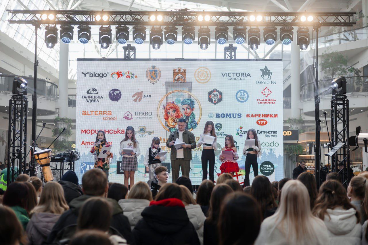 Група дітей та чоловік виступають на освітленій сцені всередині торгового центру перед аудиторією. На фоні сцени встановлено великий банер із численними логотипами партнерів та ілюстрацією по центру.