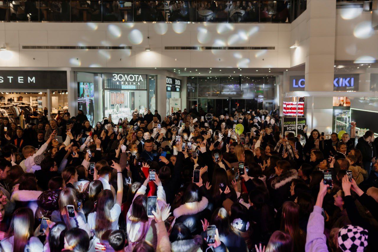 Велика кількість людей, переважно молоді, зібралася в холі торгового центру. Багато хто з присутніх тримає підняті вгору смартфони, знімаючи подію; приміщення яскраво освітлене святковими вогнями.