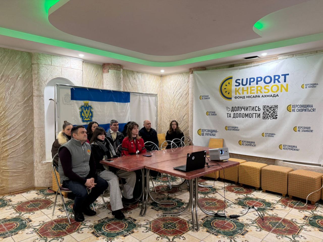 Група людей сидить за столом під час офіційної зустрічі або презентації. У приміщенні розміщено банер благодійного фонду «Support Kherson» із закликом долучитися до допомоги, а також прапор із гербом міста. На столі стоїть ноутбук та проєктор.