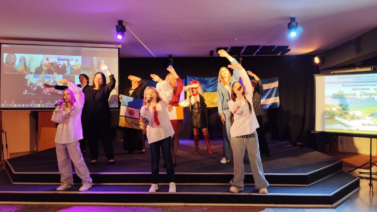 На сцені під час культурного заходу виступає група дітей та дорослих. Учасники в сучасному одязі та вишиванках співають і танцюють на фоні прапорів. З обох боків сцени встановлено екрани: на одному триває відеоконференція, на іншому — заставка фестивалю «Єдина Херсонщина».