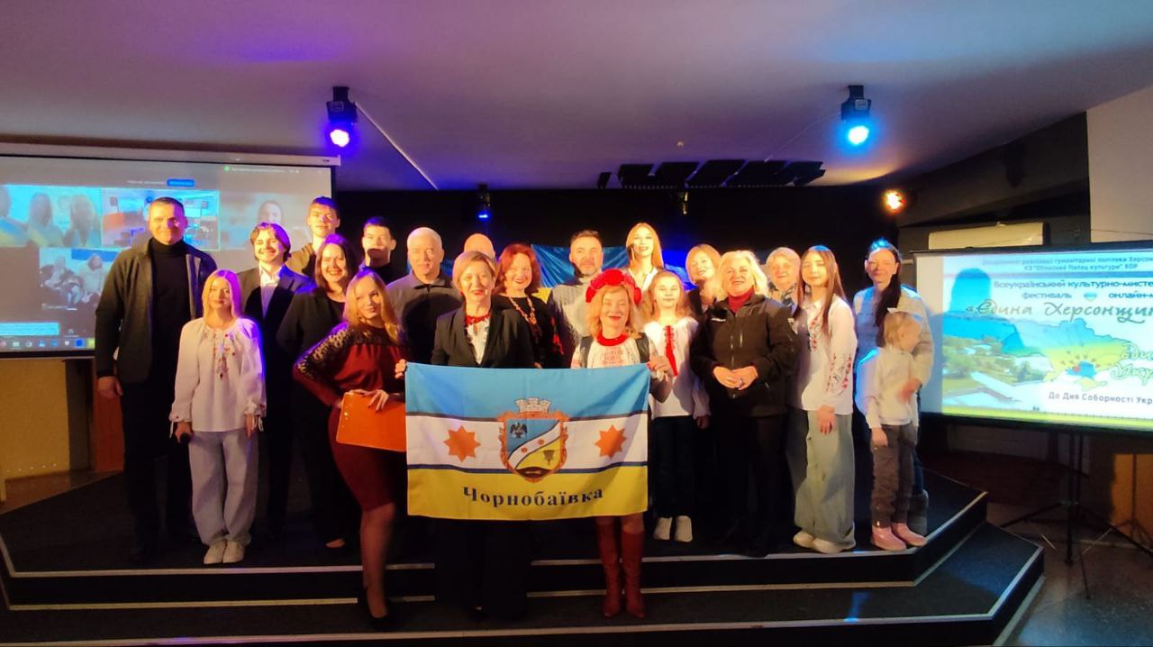 Велика група учасників культурного заходу стоїть на сцені для спільного фото. У центрі вони тримають прапор села Чорнобаївка. Позаду них розміщено державний прапор України та інші стяги, а з обох боків сцени встановлено екрани з інформацією про проєкт «Єдина Херсонщина» та вікном відеоконференції.