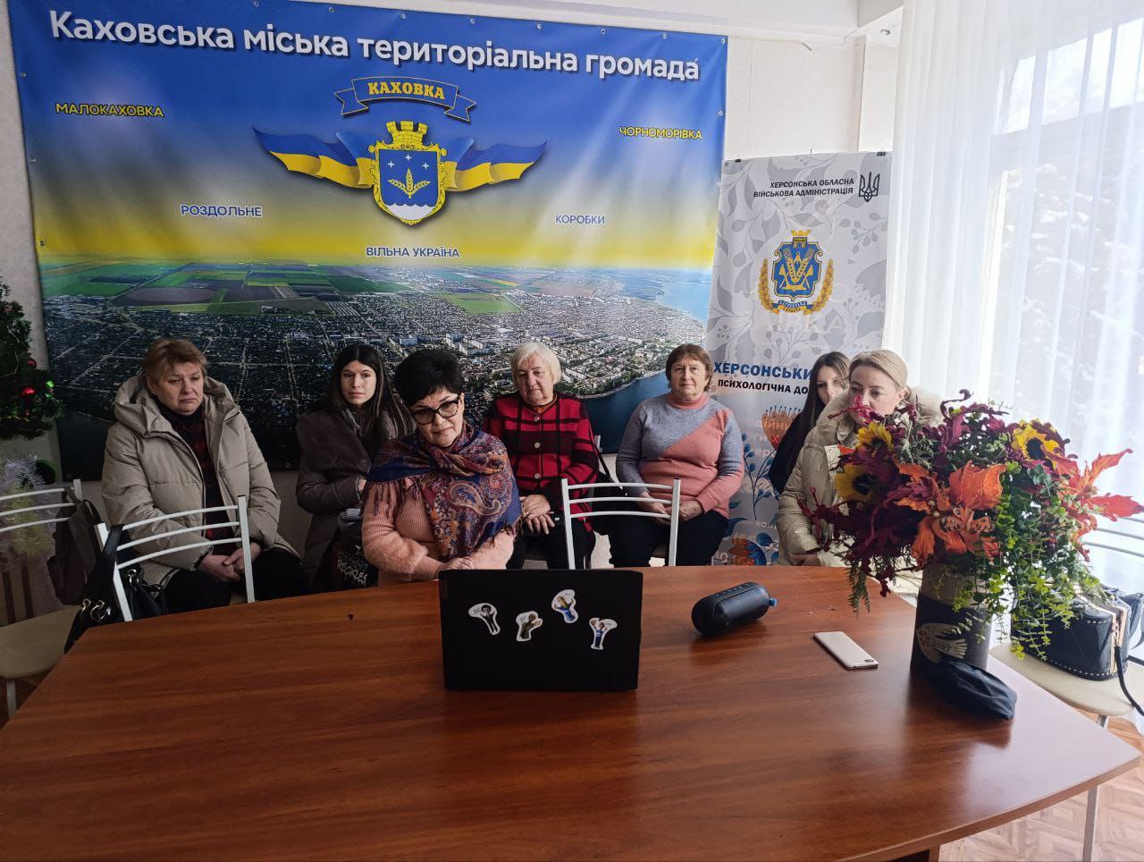Група жінок сидить за столом під час офіційного заходу на фоні великого банера Каховської міської територіальної громади. Поруч встановлено інформаційний стенд Херсонської обласної військової адміністрації. На передньому плані на столі стоїть відкритий ноутбук та ваза з осінніми квітами.