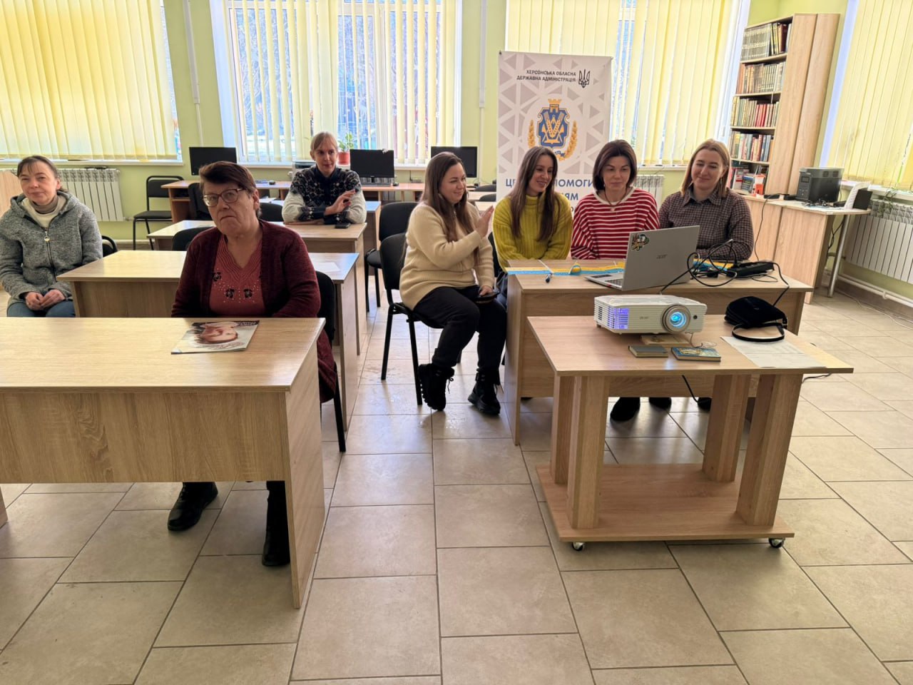 Група жінок перебуває у світлому навчальному класі або бібліотеці. Вони сидять за партами, на одній з яких стоїть ноутбук та проєктор. На задньому плані видно мобільний стенд Херсонської обласної державної адміністрації та книжкову шафу.