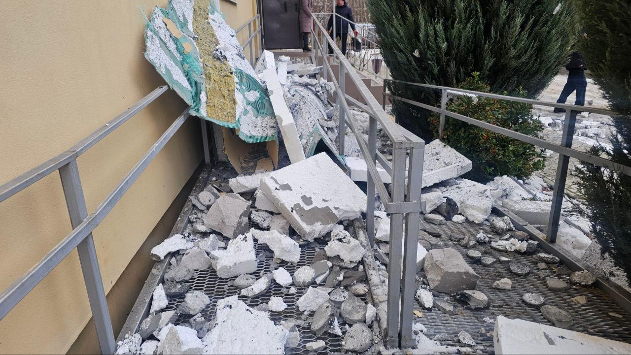 Пандус біля входу до будівлі, засипаний уламками бетону, цегли та пінопласту після обстрілу. Металеві поруччя частково пошкоджені, а на задньому плані видно вхідні двері та понівечену обшивку стіни.