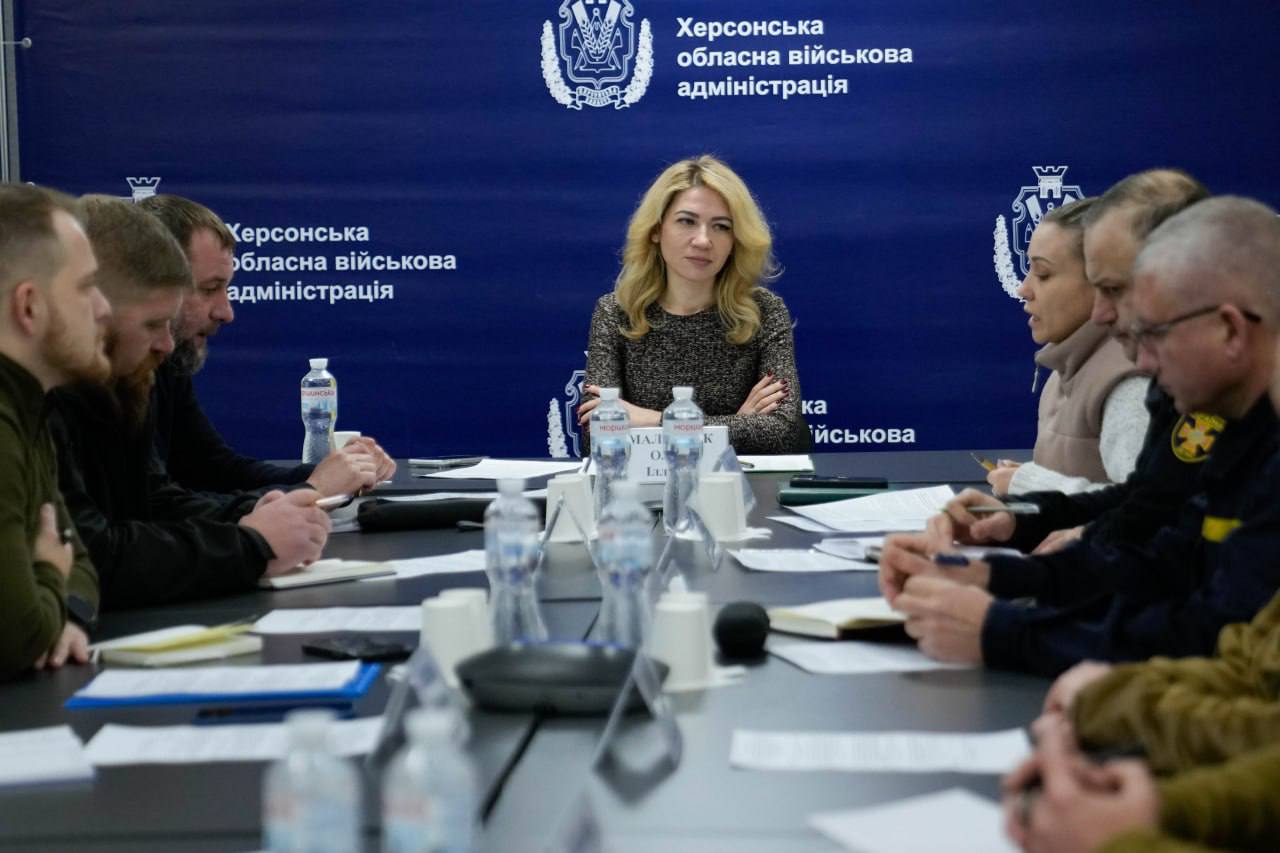 Робоча зустріч у Херсонській обласній військовій адміністрації. У центрі столу сидить жінка, яка головує на нараді, на фоні синього банера з офіційною назвою установи та гербом. Уздовж столу сидять інші учасники обговорення, перед якими лежать документи та стоять пляшки з водою.