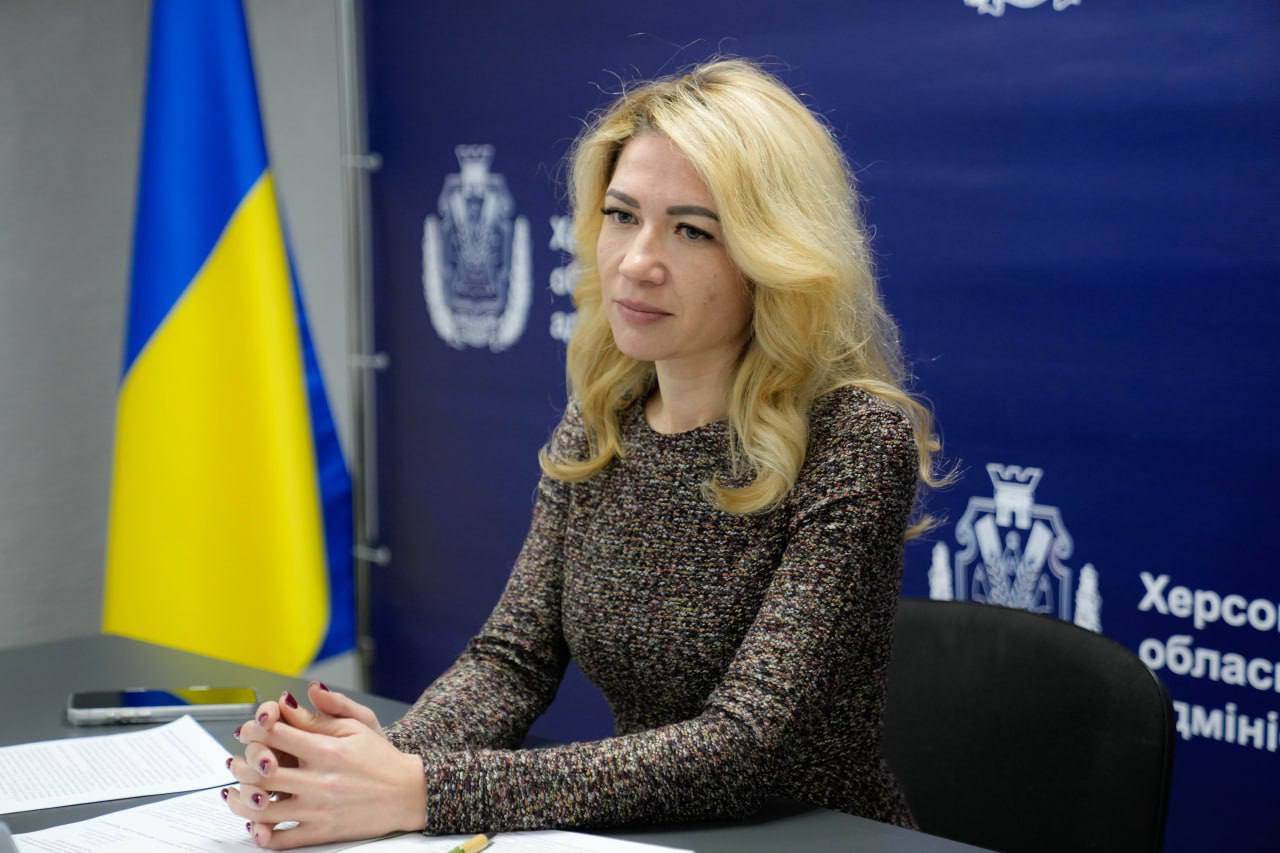 Жінка сидить за робочим столом на фоні офіційного банера Херсонської обласної військової адміністрації та державного прапора України. Перед нею лежать документи у папці, вона зосереджено дивиться вбік.