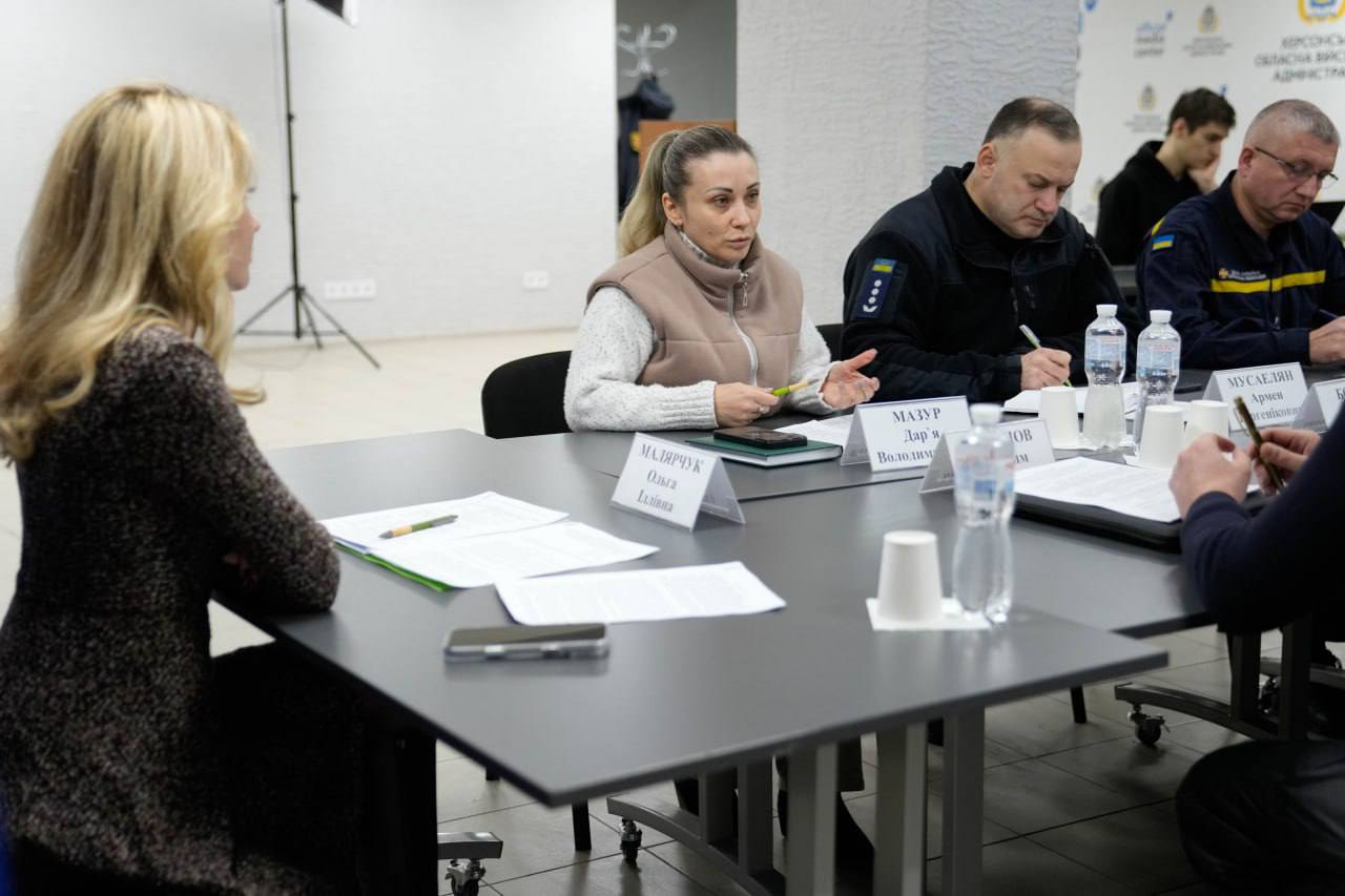 Робоча зустріч за участю представників офіційних структур та рятувальних служб. Учасники сидять за столом, на якому розміщені таблички з іменами, документи та пляшки з водою. Жінка в центрі кадру веде діалог з колегами, на фоні видно частину інформаційного стенда.