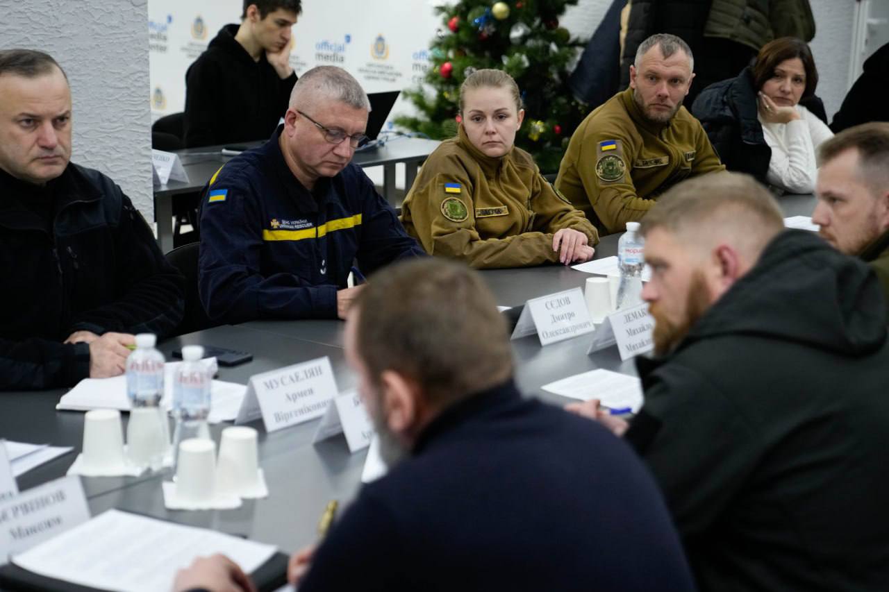 Група людей сидить за довгим столом під час робочої наради. Серед присутніх — представники ДСНС у синій формі та люди у військовій формі. На столі перед учасниками стоять таблички з іменами, пляшки з водою та лежать документи. На задньому плані помітно прикрашену ялинку.