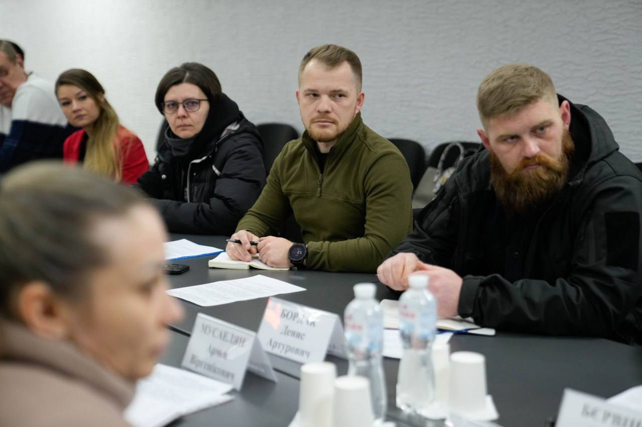 Учасники робочої наради сидять за столом, уважно слухаючи доповідача. Серед присутніх — чоловіки у тактичному одязі та цивільні особи. Перед ними на столі розміщені таблички з іменами, папери для записів та пляшки з водою.