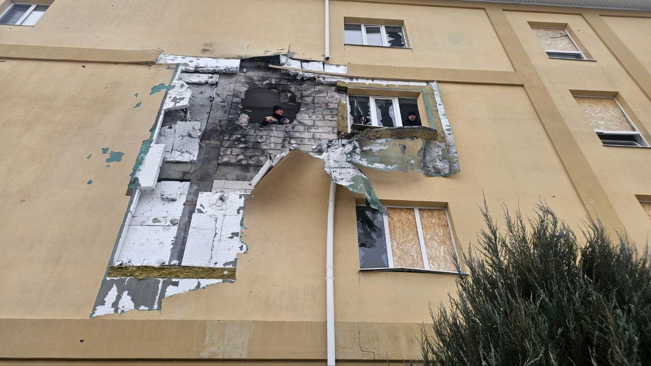 Фасад багатоповерхового будинку з великою діркою та значними руйнуваннями зовнішньої стіни внаслідок обстрілу. Видно пошкоджені балкони, вибиті вікна, деякі з яких забиті ОСБ-плитами. Крізь зруйновану стіну помітно людей усередині приміщення.