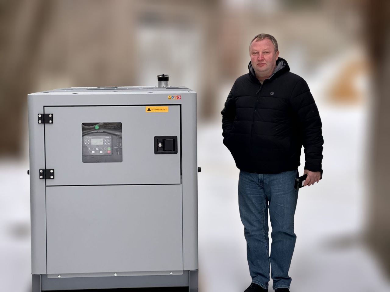 Чоловік у чорній куртці та синіх джинсах стоїть поруч із великим сірим промисловим генератором на фоні засніженого подвір'я.