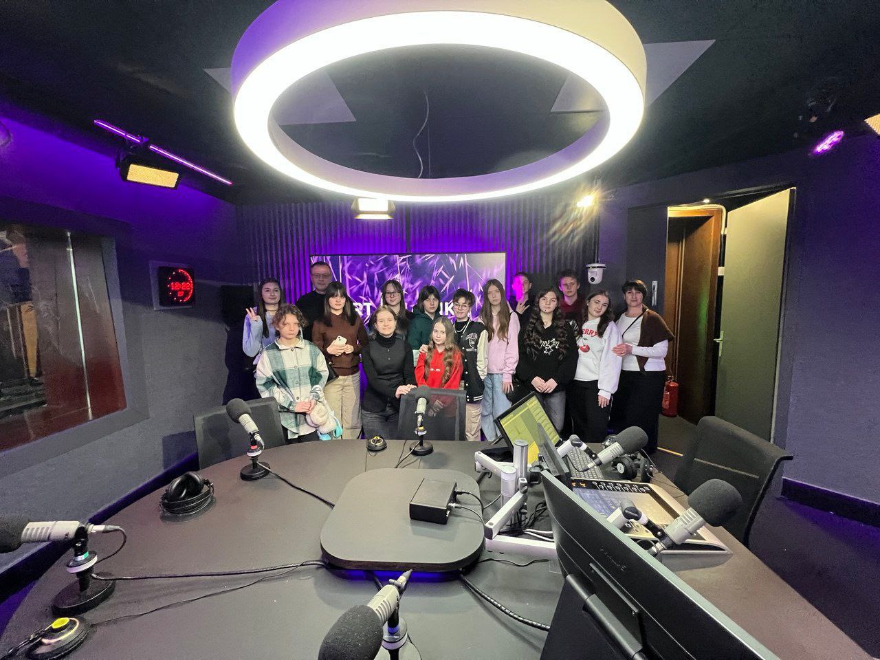 Група молоді та дорослих у сучасній студії звукозапису або радіостудії. Учасники стоять за круглим столом із мікрофонами під великим кільцевим світильником, на фоні фіолетового підсвічування та професійного обладнання.