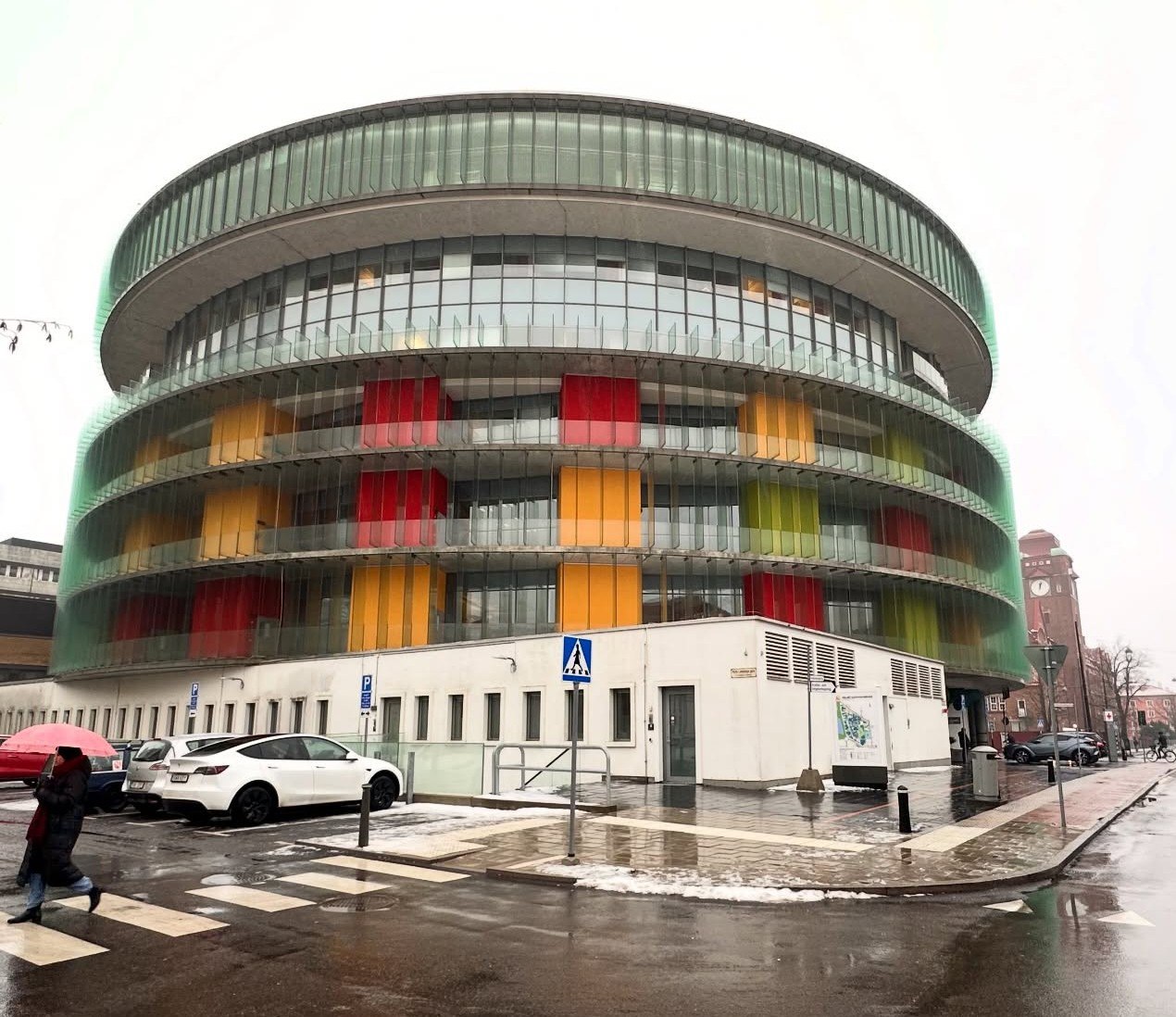 Сучасна багатоповерхова будівля круглої форми зі скляним фасадом. Фасад оздоблений вертикальними кольоровими панелями червоного, помаранчевого та зеленого кольорів. Перед будівлею розташована парковка з автомобілями та пішохідний перехід, на вулиці лежить трохи снігу.