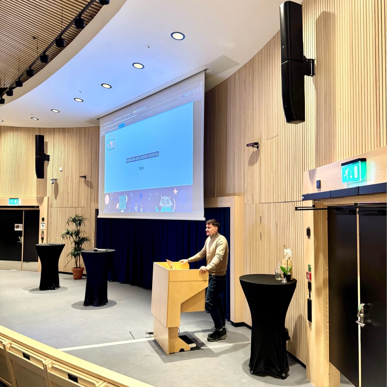 Чоловік стоїть за трибуною у великій сучасній конференц-залі з дерев'яним оздобленням стін. На великому екрані над сценою відображається презентація з написом «KHERSON CHILDREN'S HOSPITAL 2026». У залі також видно високі коктейльні столи, накриті чорною тканиною.