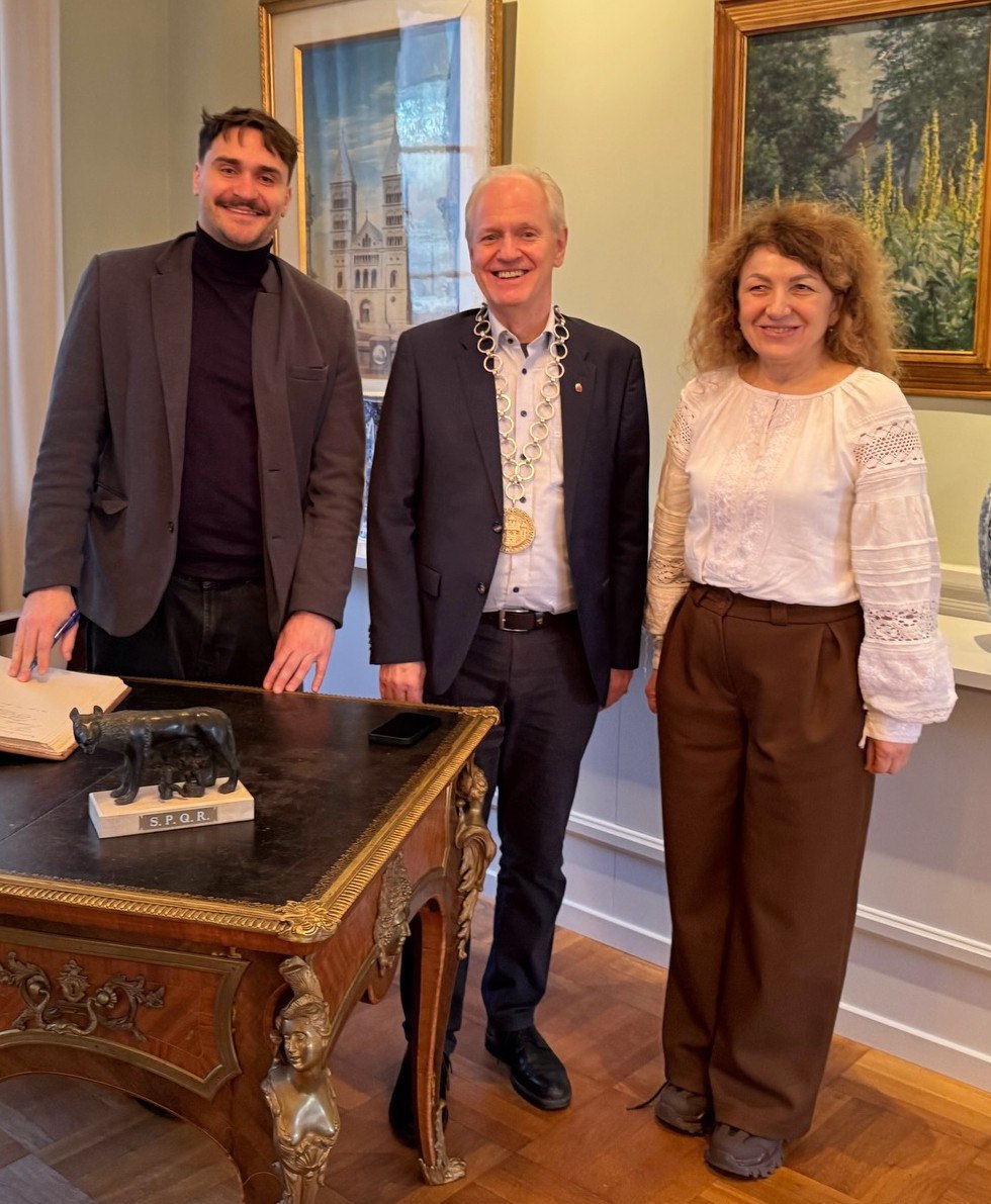 Троє людей — двоє чоловіків та жінка — стоять поруч у класичному кабінеті. Чоловік по центру вдягнений у костюм з масивною золотистою церемоніальною вервицею на шиї, жінка — у білу вишиванку та коричневі штани. Поряд розташований антикварний стіл із відкритою книгою та статуеткою капітолійської вовчиці.