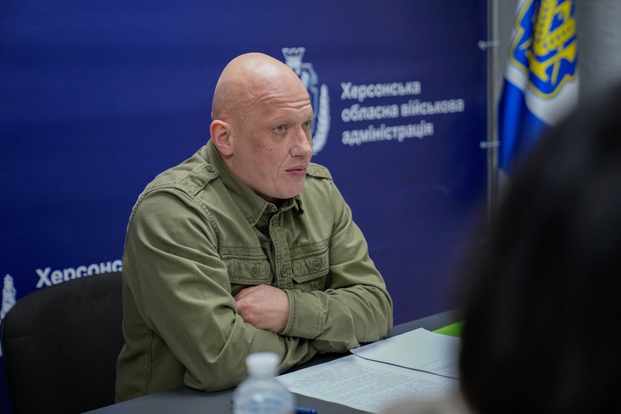 Портрет чоловіка у військовій сорочці оливкового кольору, який сидить за столом із паперами під час наради. Він уважно слухає співрозмовника, склавши руки перед собою. На фоні — синій банер із написом «Херсонська обласна військова адміністрація» та державна символіка.