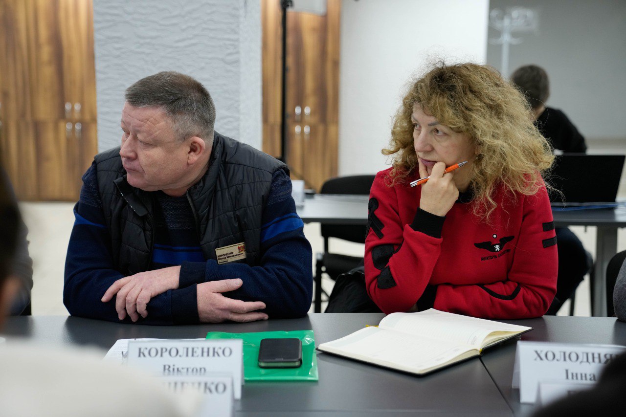 Учасники робочої наради в офісному приміщенні сидять за столом. Чоловік у темному жилеті дивиться вбік, поруч жінка у червоному світшоті робить нотатки в блокноті. Перед ними на столі стоять таблички з іменами.