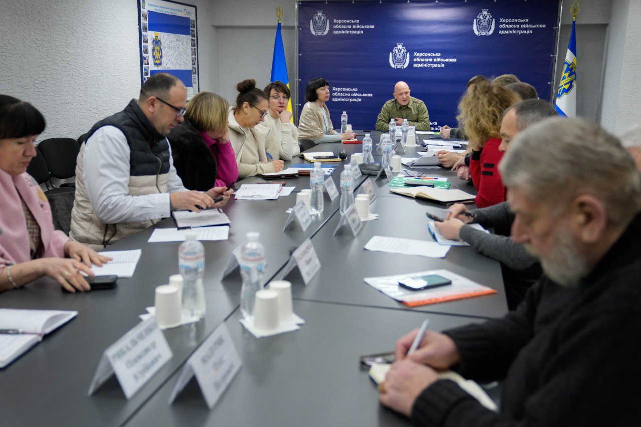 Робоча нарада в Херсонській обласній військовій адміністрації. Група людей сидить за довгим столом під час обговорення, на фоні — синій банер із гербом та прапор України. Чоловік у військовій формі на чолі столу веде зустріч.