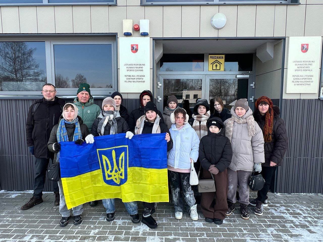 Група підлітків та дорослих у зимовому одязі стоїть перед входом до будівлі в Литві. Учасники тримають перед собою розгорнутий державний прапор України з гербом. На фасаді будівлі розміщені таблички литовською мовою та знак укриття.