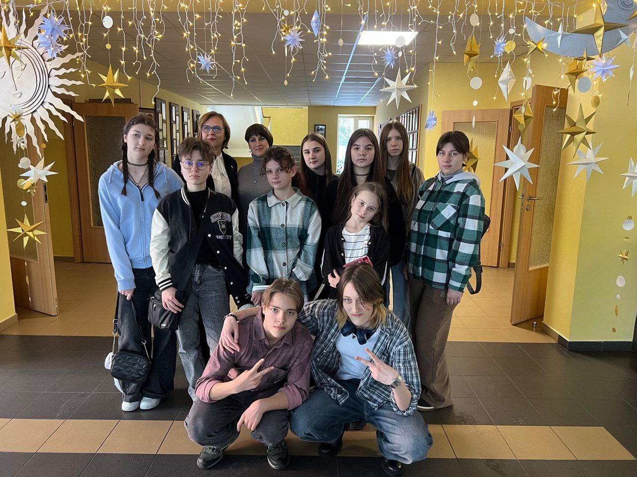 Група підлітків та двоє дорослих позирують у світлому коридорі навчального закладу. Приміщення прикрашене декоративними підвісними зірками, місяцем та гірляндами. Двоє хлопців на передньому плані присіли та показують жест «мир», інші учасники стоять позаду.