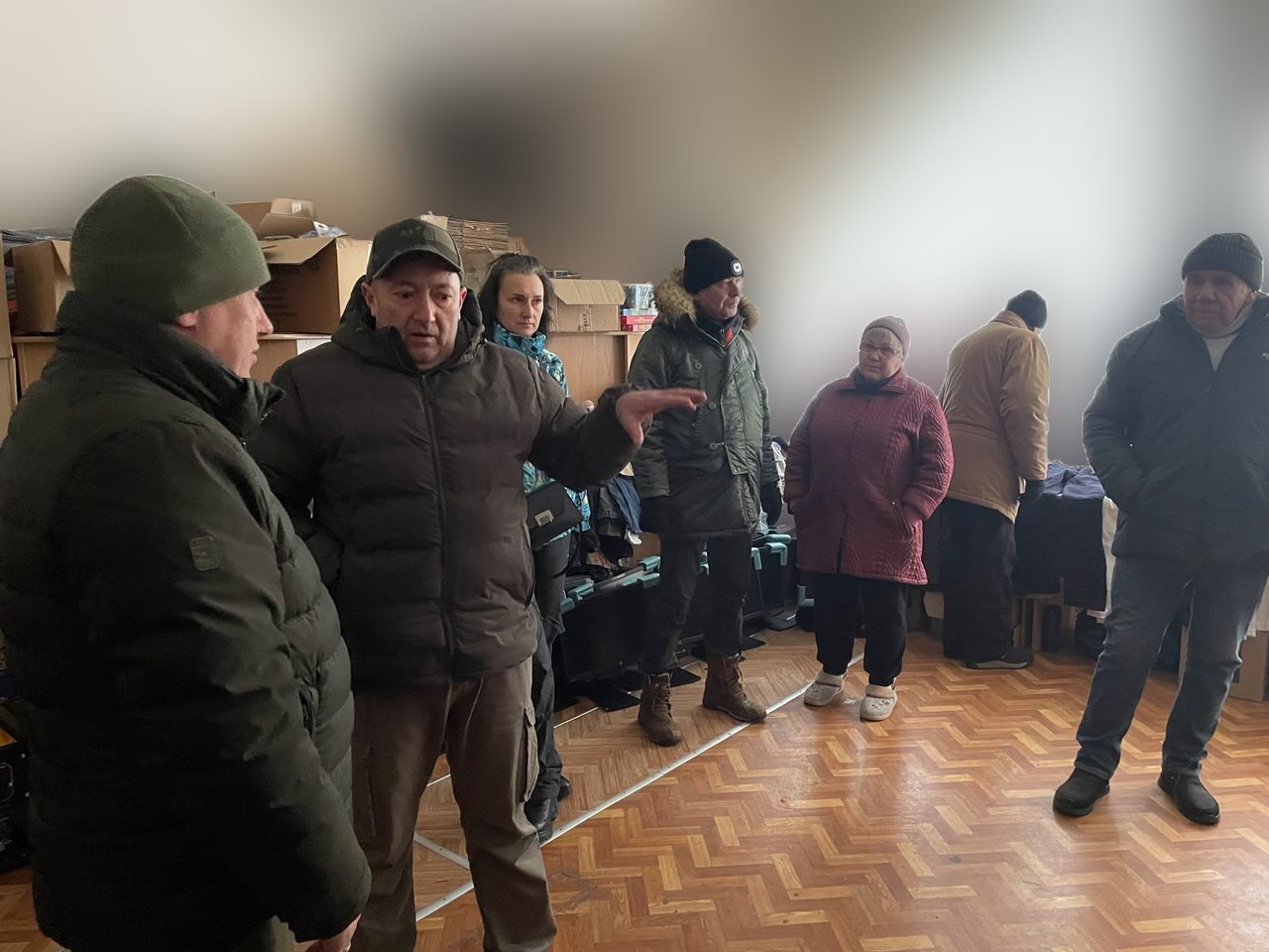 Група людей у зимовому одязі зібралася в приміщенні складу поруч із пакунками та речами.