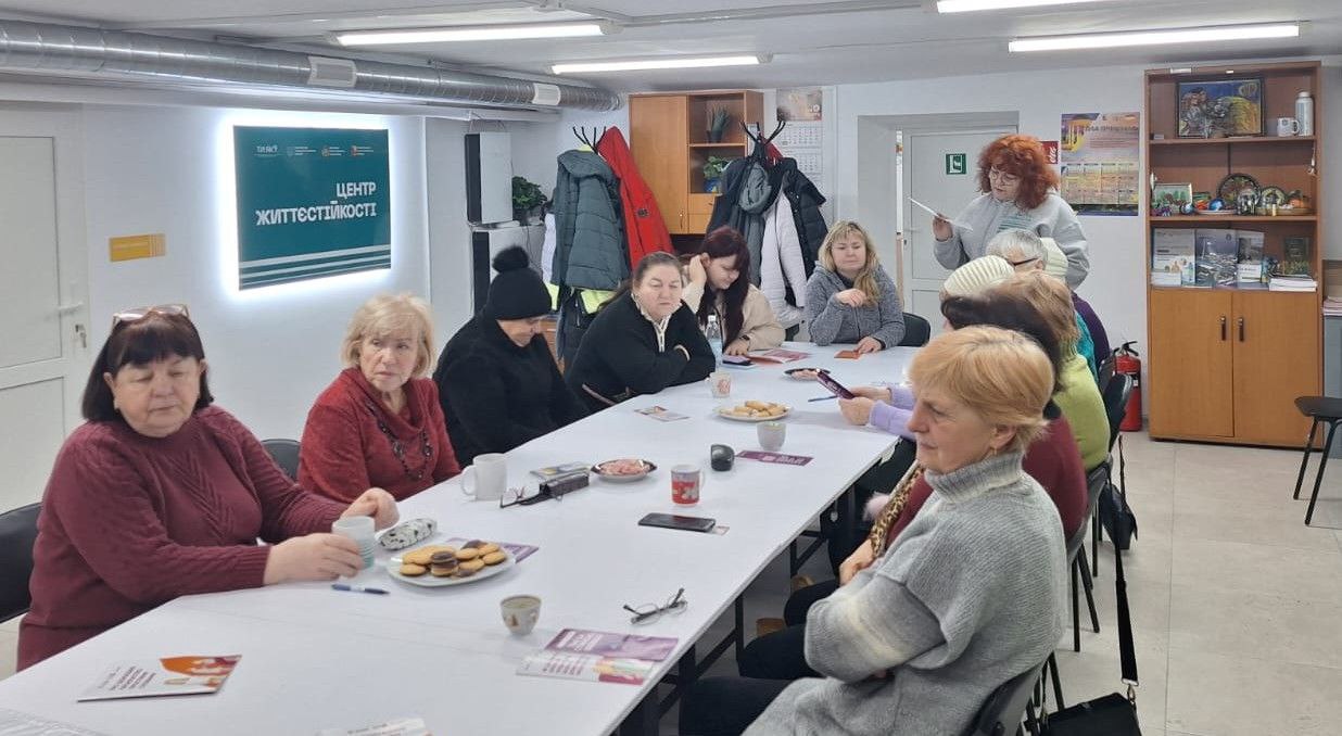 Група жінок за довгим столом у світлому приміщенні «Центру життєстійкості». Учасниці спілкуються, на столі стоять чашки та частування. На задньому плані видно вішалку з одягом, шафу та світлову вивіску з назвою закладу.