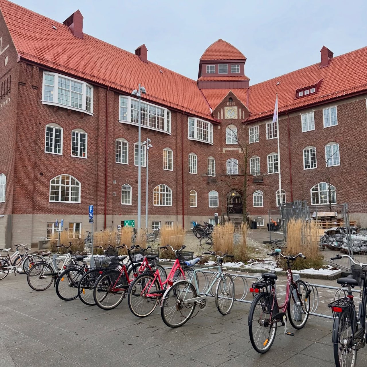 Велика триповерхова цегляна будівля з червоним черепичним дахом та вежею з годинником у центрі фасаду. Перед будівлею розташована велика велопарковка з різнокольоровими велосипедами, а на подвір’ї видно залишки снігу та декоративні кущі сухої трави.