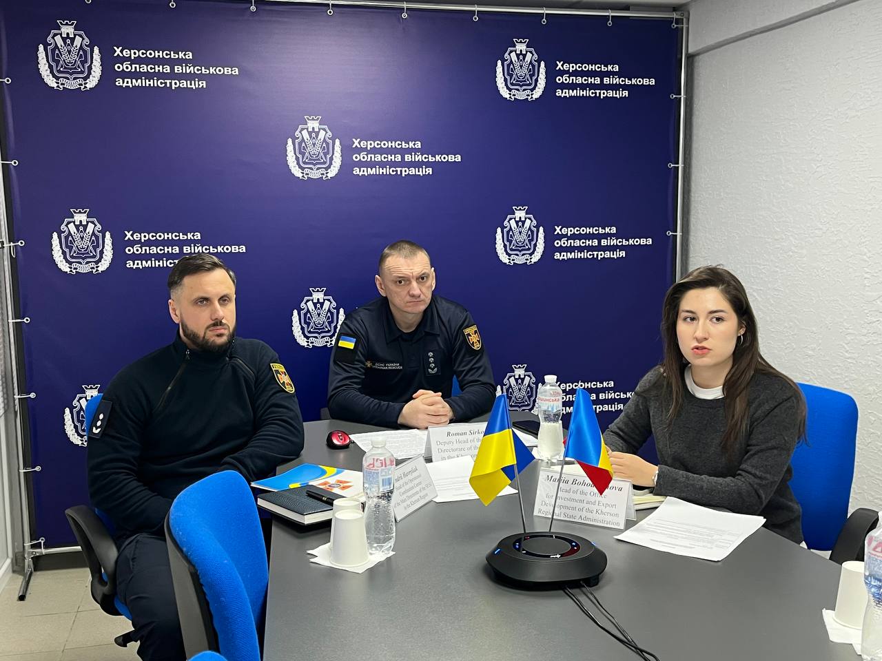 Двоє чоловіків у чорній формі з шевронами та жінка у цивільному одязі сидять за робочим столом під час наради на фоні банера Херсонської ОВА з українською символікою.