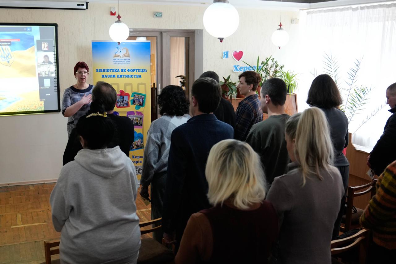 Учасники заходу в бібліотеці стоять під час прослуховування промови або гімну. Попереду стоїть жінка-доповідач, а поруч із нею розміщено інформаційний стенд «Бібліотека як фортеця: пазли дитинства».