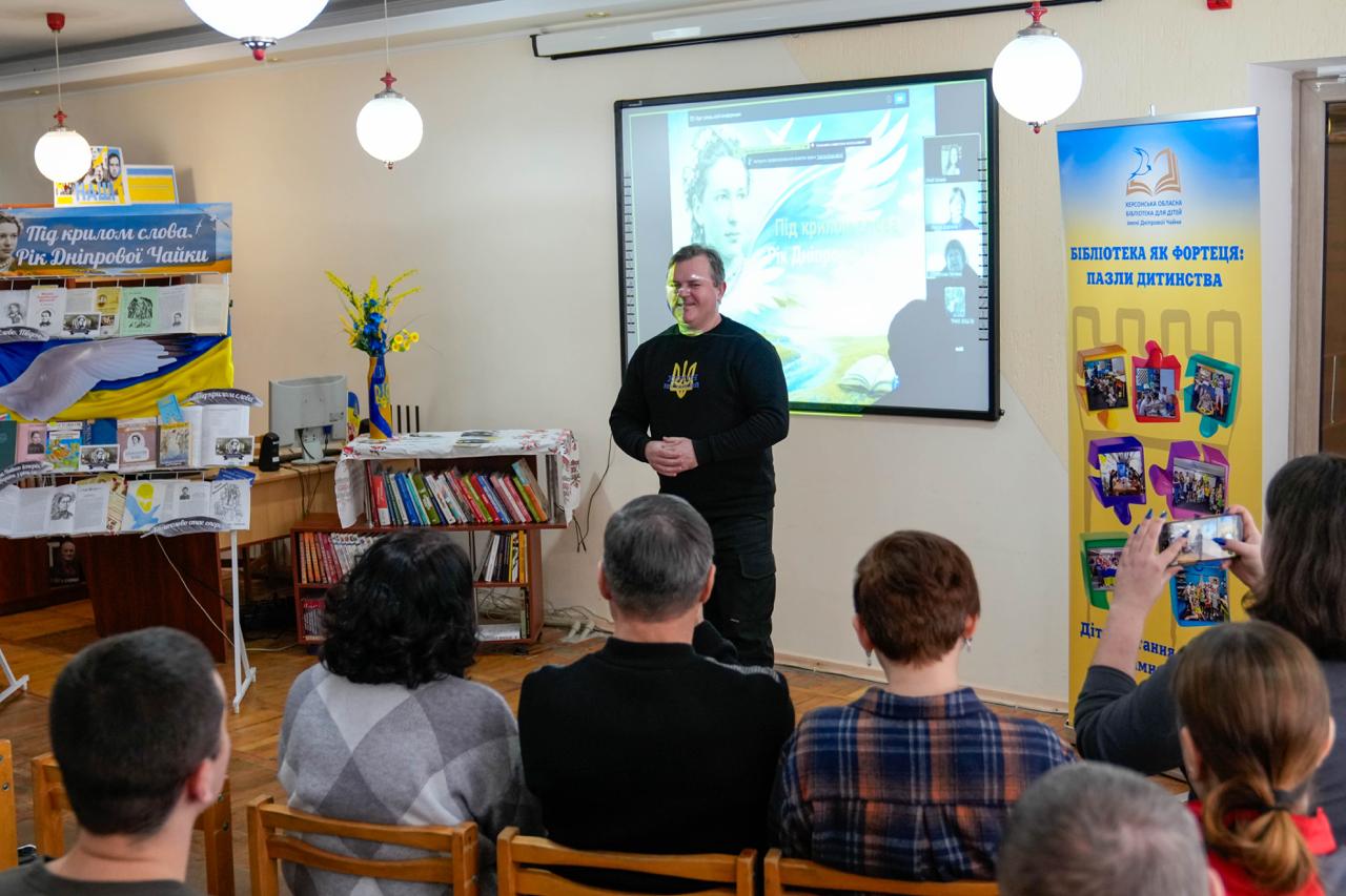 У приміщенні бібліотеки проходить презентація за участю слухачів та доповідача, який стоїть біля екрана з проєкцією. У залі розміщено тематичні банери та книжкову виставку «Під крилом слова. Рік Дніпрової Чайки».