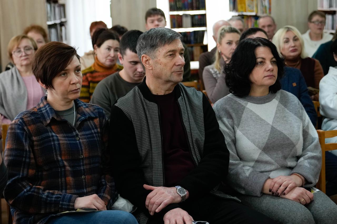 Глядачі в залі бібліотеки уважно слухають виступ під час презентації. На передньому плані сидять чоловік та дві жінки, на фоні видно інші ряди з відвідувачами та книжкові полиці.