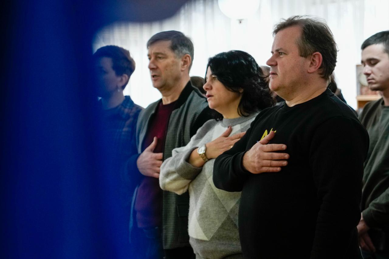 Учасники заходу в бібліотеці стоять, приклавши праву руку до серця під час урочистої миті. На фоні видно книжкові полиці, а на передньому плані — чоловіків та жінку в офіційно-казуальному одязі.