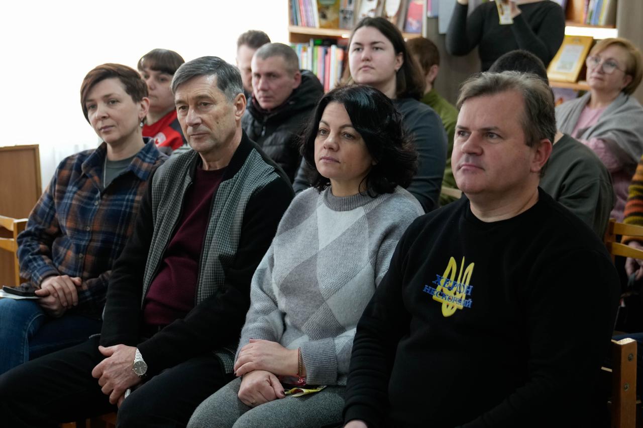 Група людей у залі бібліотеки сидить на стільцях під час офіційного заходу. На передньому плані видно чоловіка у чорному світшоті з написом «Херсон нескорений» та тризубом, а поруч із ним — інших зосереджених слухачів.