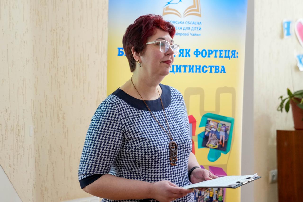 На світлині жінка-спікер у сукні з дрібним візерунком та окулярах виступає перед аудиторією. Вона тримає планшет із текстом промови. Поруч розміщено банер «Бібліотека як фортеця: пазли дитинства» із логотипом Херсонської обласної бібліотеки для дітей імені Дніпрової Чайки.