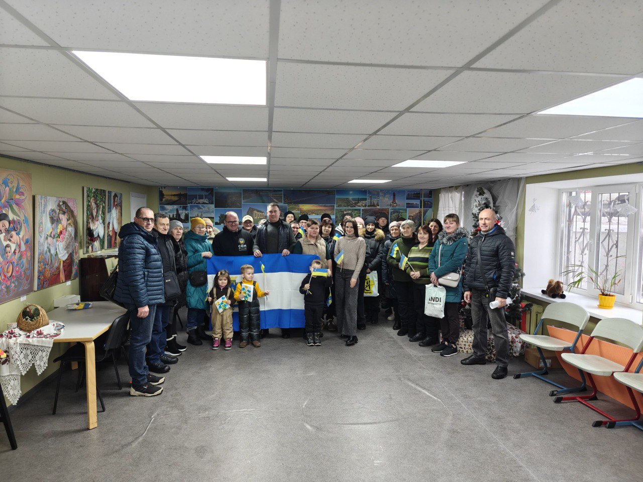 Велика група людей різного віку, серед яких є маленькі діти, позує для спільного фото у світлому приміщенні. У центрі вони тримають великий синьо-білий прапор, а діти тримають у руках маленькі прапорці. Стіни кімнати прикрашені картинами та фотографіями.