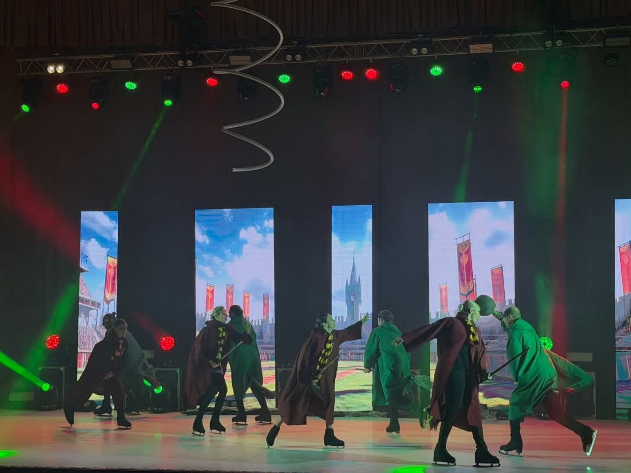 Фігуристи на льодовій арені виконують театралізований номер у довгих мантіях, що нагадують стилістику «Гаррі Поттера». На фоні встановлено високі екрани з анімацією казкового замку та яскраве сценічне освітлення.