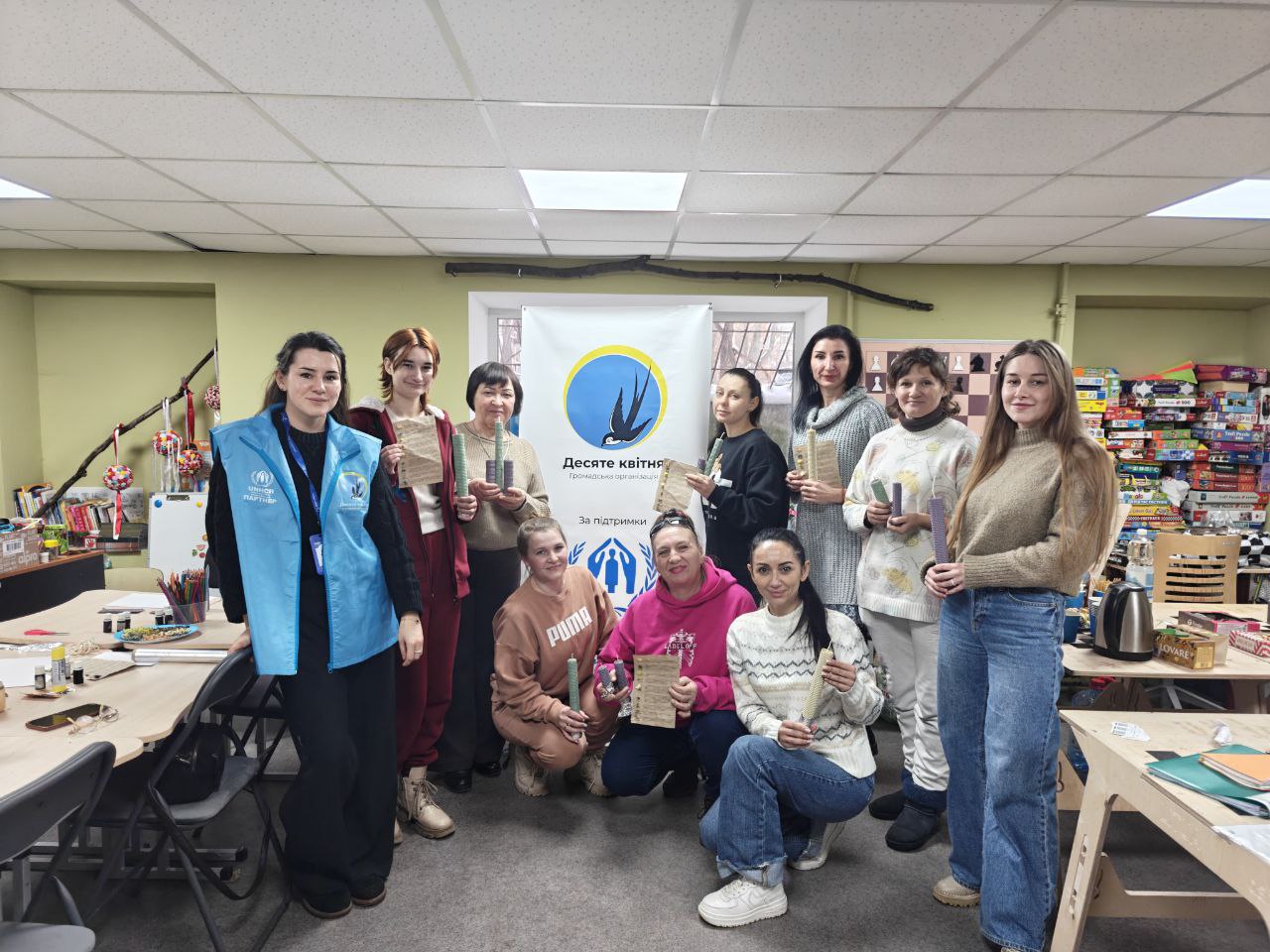 Група жінок у світлому навчальному класі позує на фоні білого банера з логотипом громадської організації. Більшість тримає в руках невеликі вироби ручної роботи, схожі на свічки або паперові сувої. На полицях праворуч видно багато настільних ігор та книг.