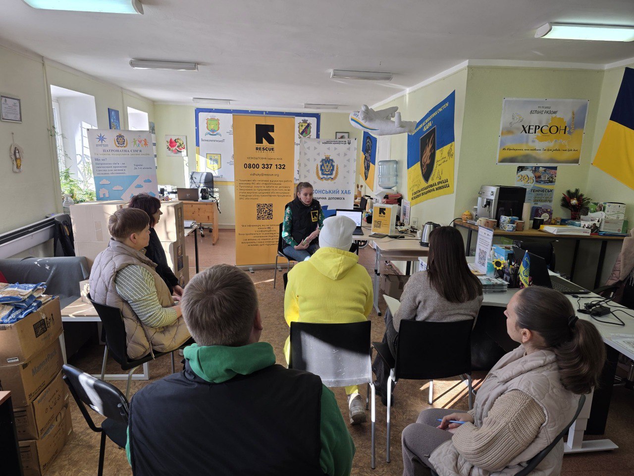 Група людей сидить півколом у світлому офісному приміщенні та слухає доповідачку, яка сидить навпроти них біля ноутбука. Навколо розміщені інформаційні банери з логотипами та контактами, державна символіка та коробки з гуманітарною допомогою.