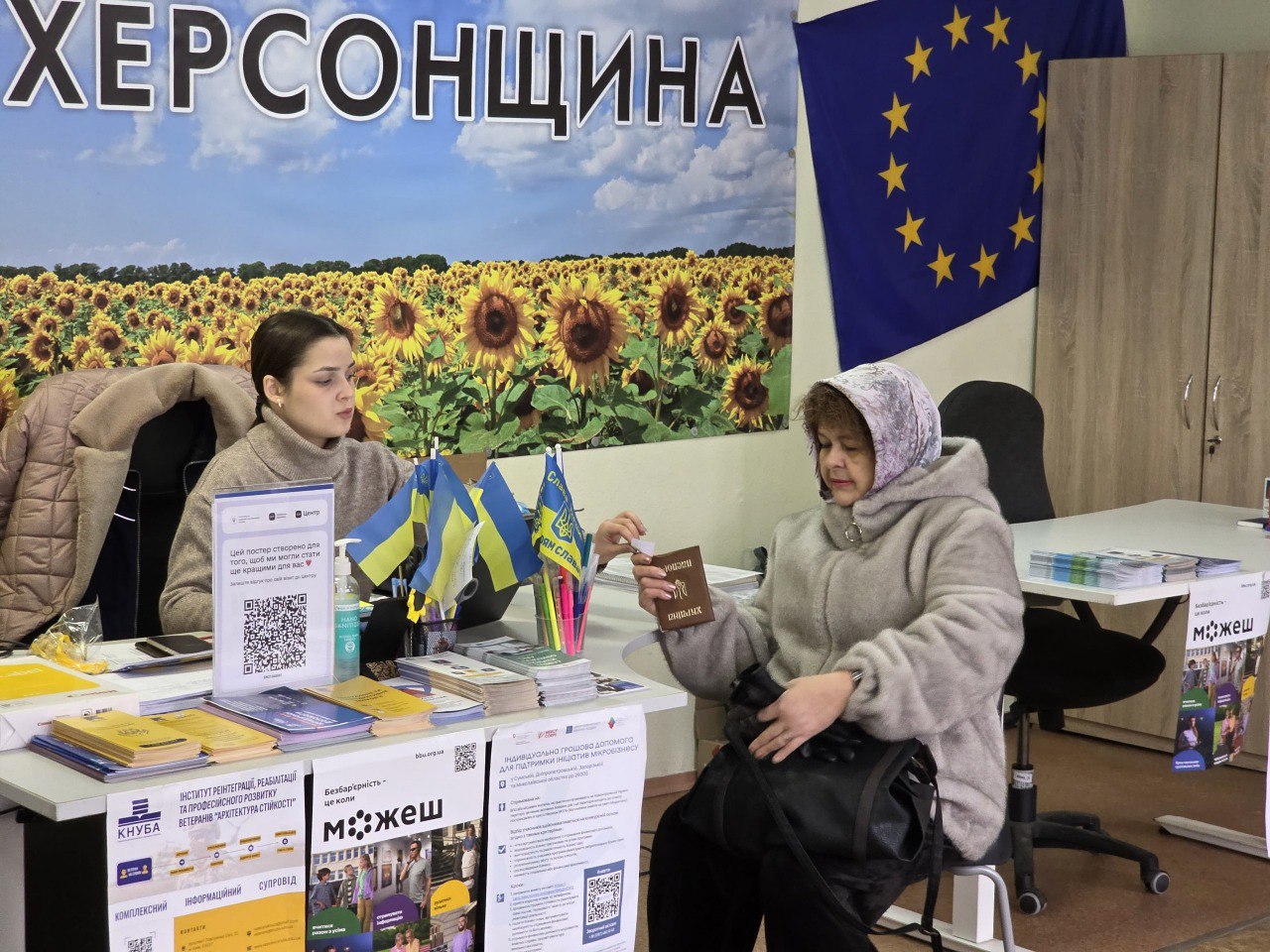 Дві жінки спілкуються за робочим столом, на якому розміщено інформаційні буклети та маленькі прапорці України. На фоні — велике панорамне фото соняшникового поля з написом «Херсонщина» та прапор Європейського Союзу на стіні.