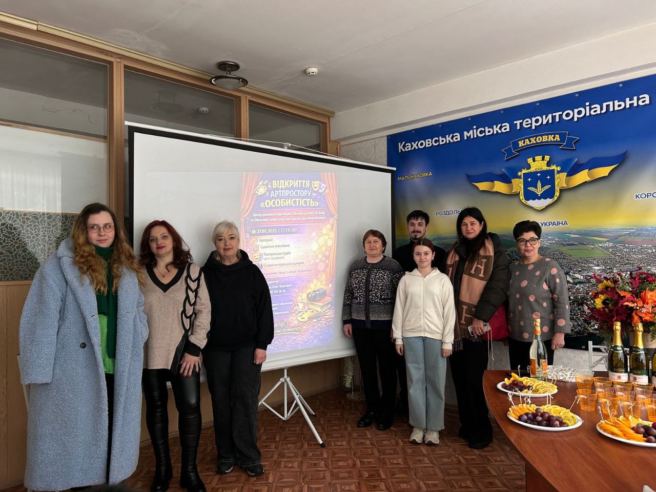 Група людей стоїть у приміщенні біля великого екрана з афішою про відкриття артпростору. Поруч на стіні розміщено банер із гербом Каховки, а на передньому плані стоїть стіл із фруктовою нарізкою та напоями.