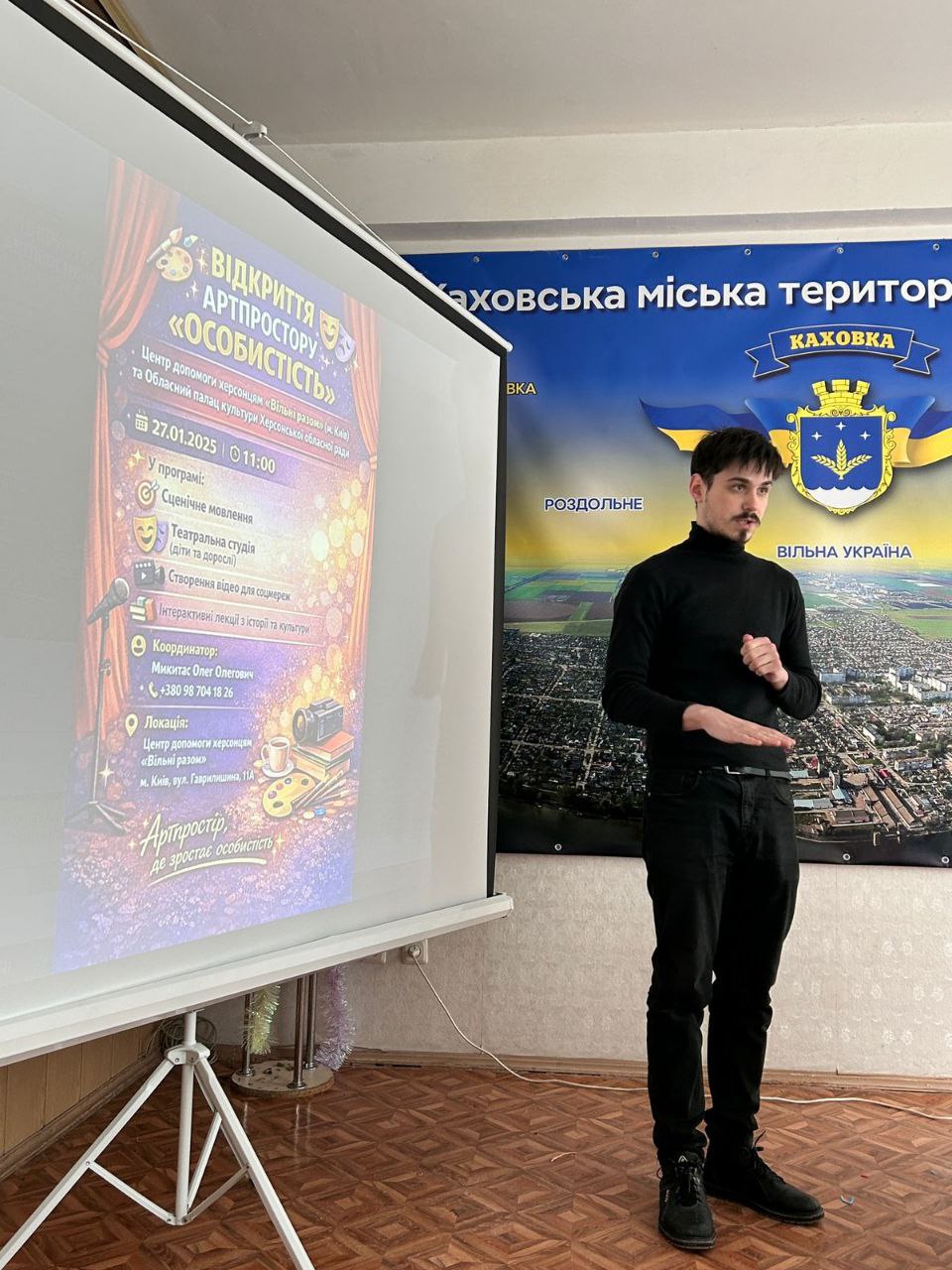 Чоловік у чорному одязі виступає з промовою перед білим екраном, на який виведено яскраву афішу про відкриття артпростору. Праворуч на стіні розміщено великий синій банер із гербом міста та написом «Каховська міська територіальна громада».